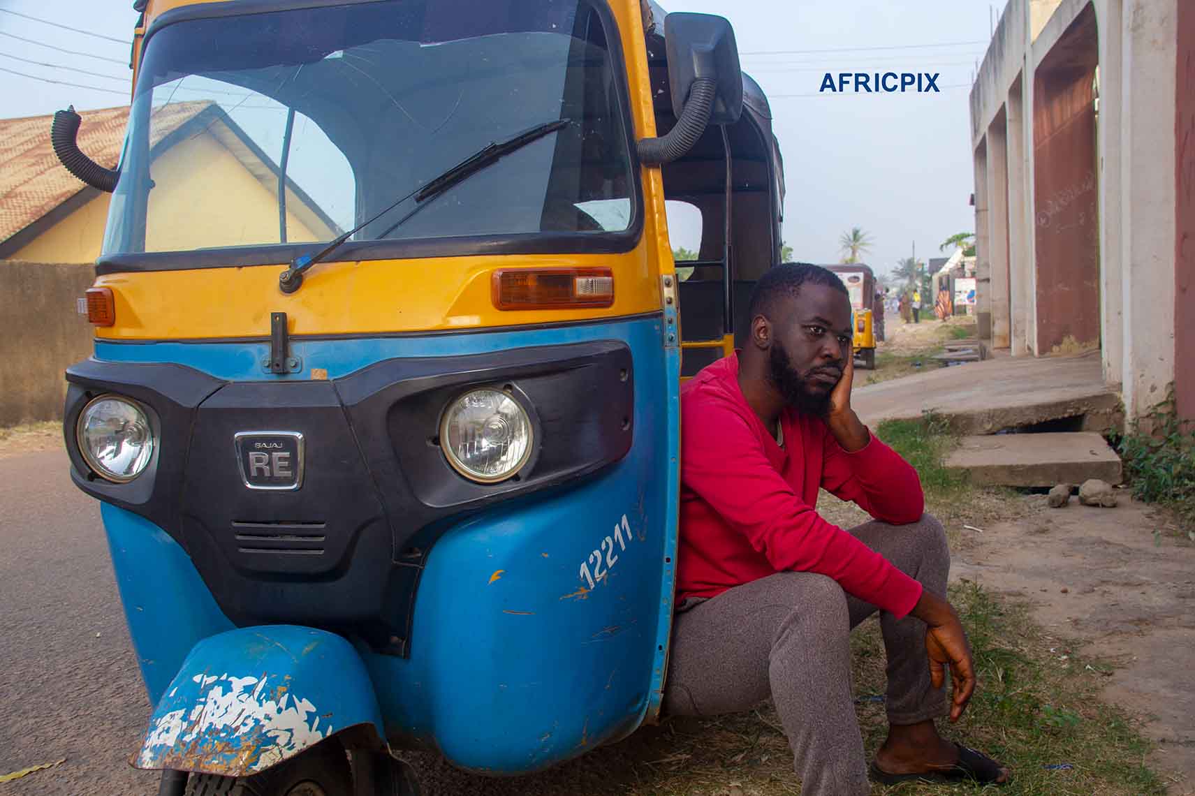 A worried African tricycle keke maruwa rider standing in front of his tricycle, with both hands on his cheeks, looking sad side view